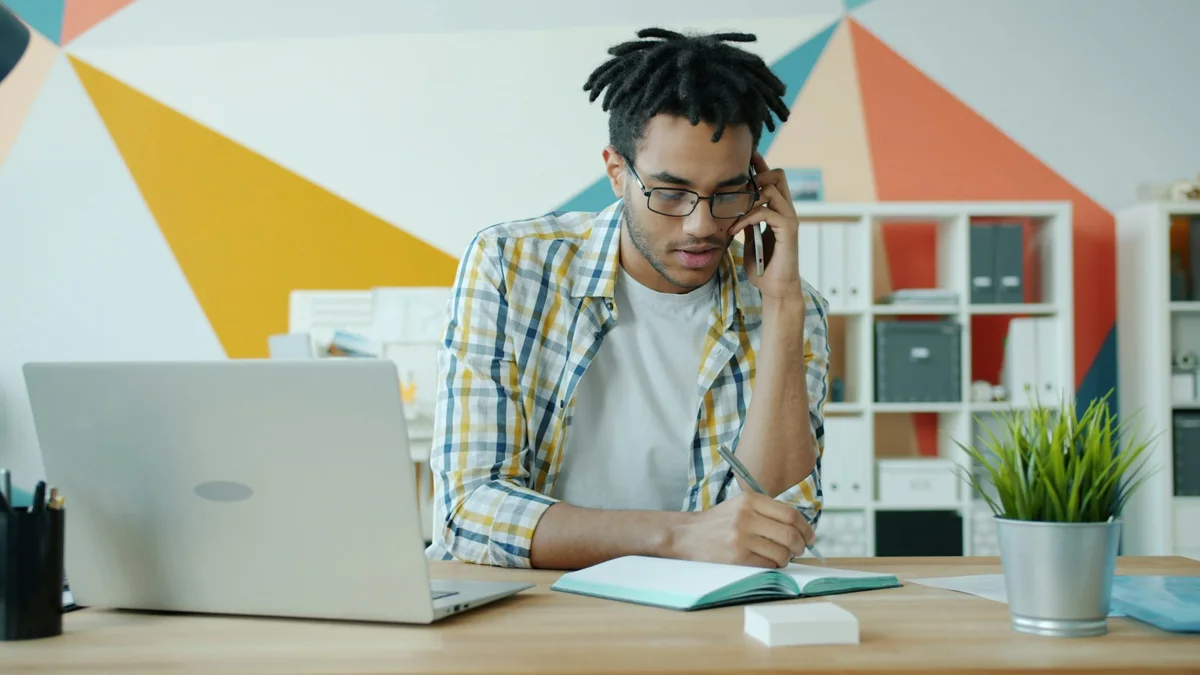 A man taking notes during a phone call beside a laptop.